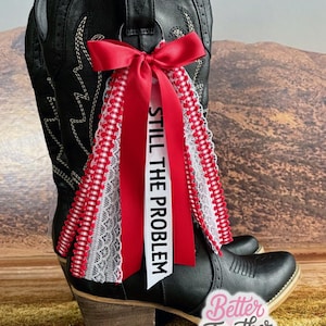 May include: Black cowboy boot with a red bow and ribbons. The ribbons are accented with white lace and a red and white gingham pattern. The text "STILL THE PROBLEM" is printed on a white ribbon.