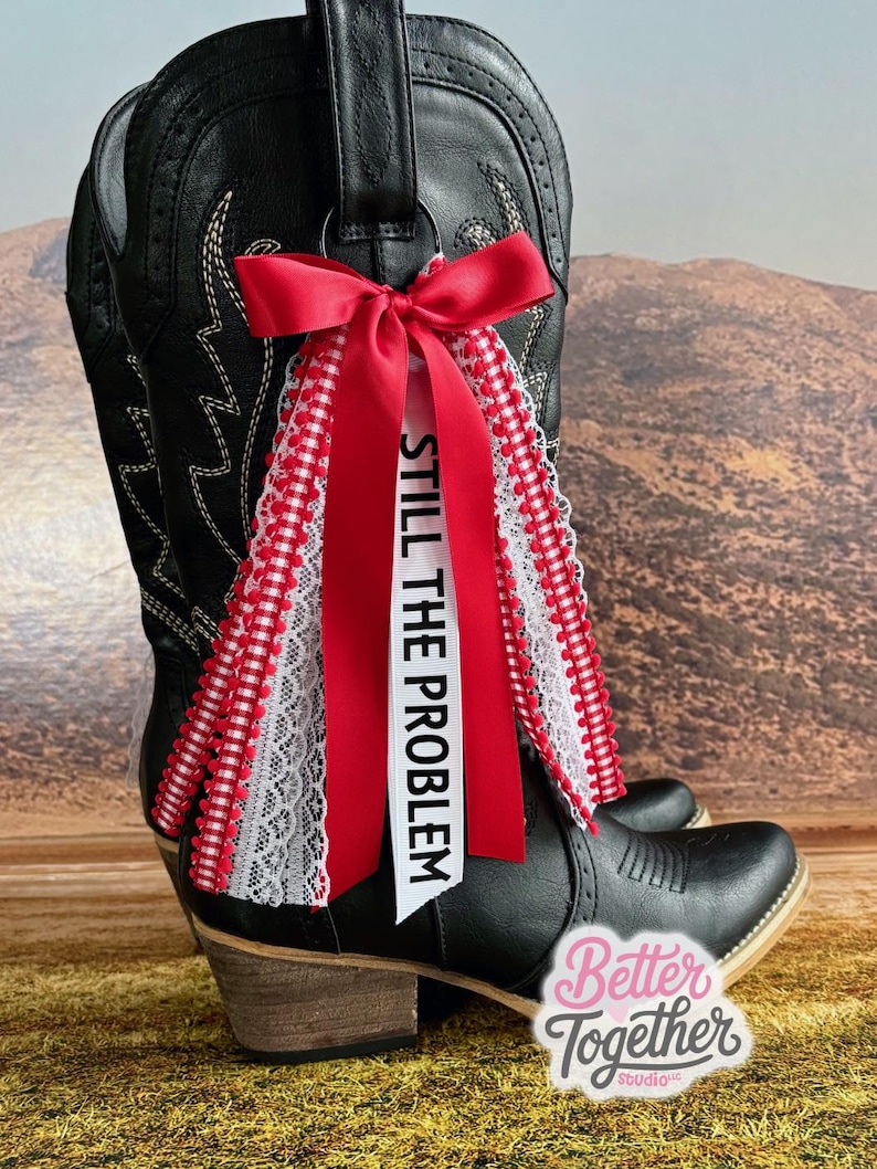 May include: Black cowboy boot with a red bow and ribbons. The ribbons are accented with white lace and a red and white gingham pattern. The text "STILL THE PROBLEM" is printed on a white ribbon.