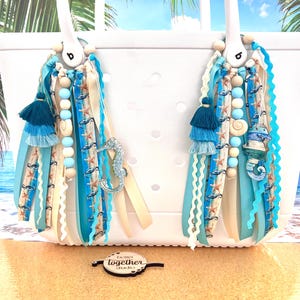 May include: Two blue and white beach bag charms with seahorse, starfish, and seashell accents. The charms are made with ribbons, beads, and tassels. The charms are attached to a white tote bag. The charms are labeled "Do it Together Studio".