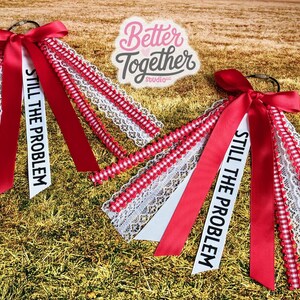 May include: Two red and white accessories with bows and ribbons. Each has a white ribbon with the text "STILL THE PROBLEM". The accessories are on a bed of dry grass. The logo "Better Together Studio" is in the background.