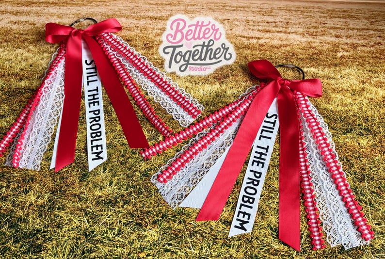 May include: Two red and white accessories with bows and ribbons. Each has a white ribbon with the text "STILL THE PROBLEM". The accessories are on a bed of dry grass. The logo "Better Together Studio" is in the background.