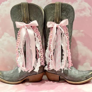 May include: A pair of distressed grey leather cowboy boots featuring decorative bows made of light pink satin ribbon, white ruffled fabric, and pink crocheted trim attached to the upper shaft of each boot, with a pointed toe and low heel.