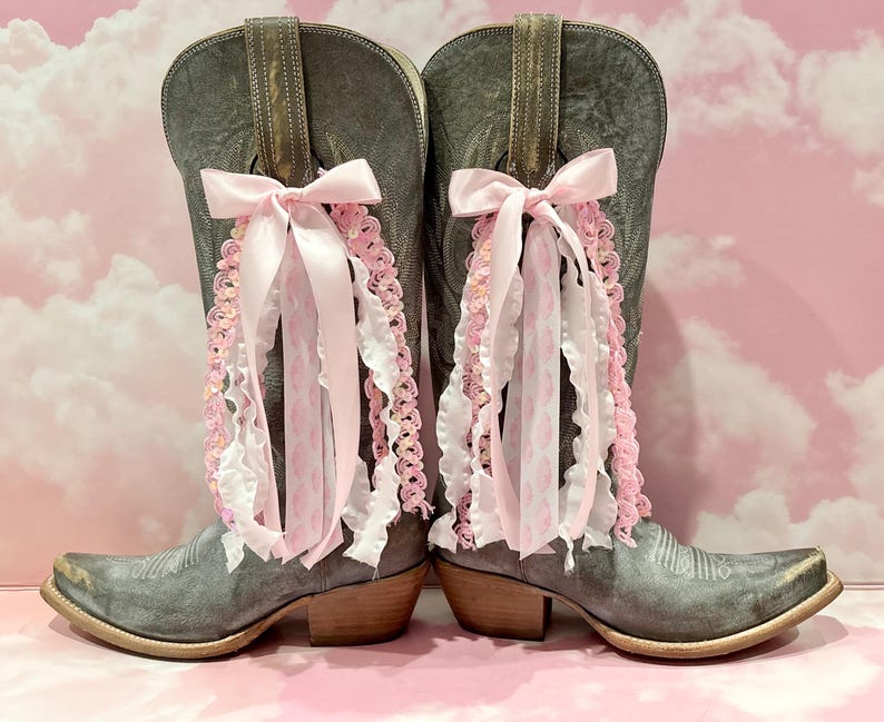 May include: A pair of distressed grey leather cowboy boots featuring decorative bows made of light pink satin ribbon, white ruffled fabric, and pink crocheted trim attached to the upper shaft of each boot, with a pointed toe and low heel.