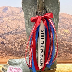 May include: A gray cowboy boot with a red, white, and blue ribbon and lace boot bling. The white ribbon says "STILL THE PROBLEM" in black text. The boot has a decorative design and a brown leather pull strap.