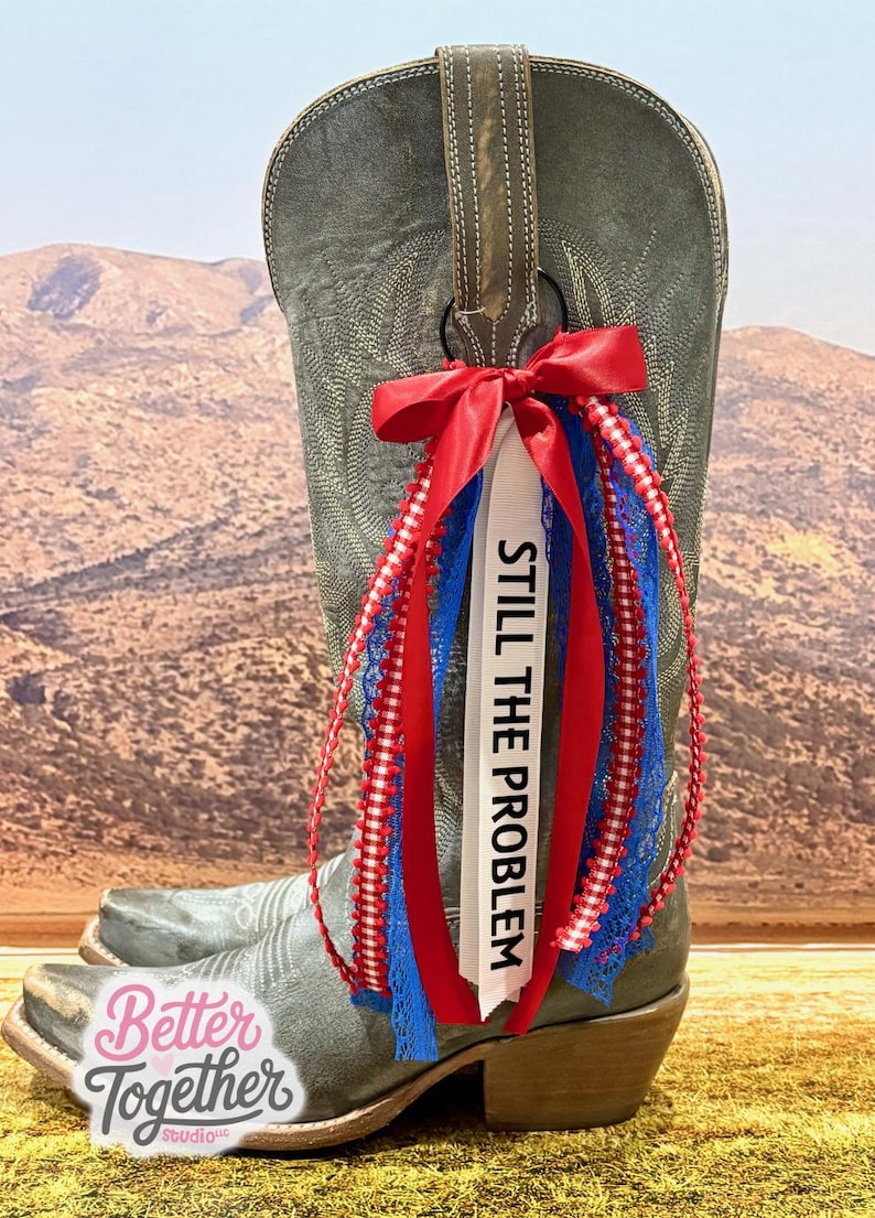 May include: A gray cowboy boot with a red, white, and blue ribbon and lace boot bling. The white ribbon says "STILL THE PROBLEM" in black text. The boot has a decorative design and a brown leather pull strap.