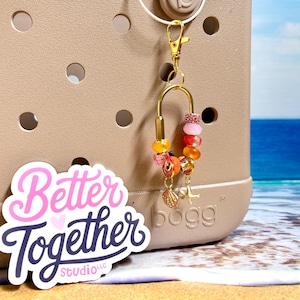 May include: A gold-toned metal keychain charm featuring a U-shaped clasp adorned with a variety of colorful beads in shades of pink, orange, and yellow. Two small dangling charms, a seashell and a starfish, hang from the bottom of the bead arrangement.