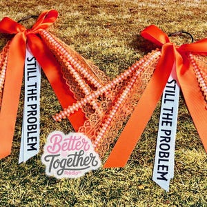 May include: Two orange hair accessories with bows, lace, and beaded accents. Each has a white ribbon printed with "STILL THE PROBLEM." A "Better Together Studio" logo is also visible.