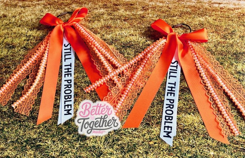 May include: Two orange hair accessories with bows, lace, and beaded accents. Each has a white ribbon printed with "STILL THE PROBLEM." A "Better Together Studio" logo is also visible.