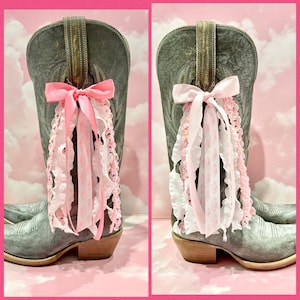 May include: A pair of grey distressed leather cowboy boots featuring decorative pull-on straps adorned with layered pink and white ribbons, including satin bows, ruffled lace trim, and patterned fabric streamers hanging from the sides.