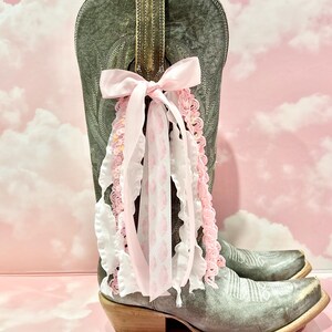 May include: A distressed grey leather cowboy boot featuring a decorative pull-on strap adorned with a multi-layered bow. The bow consists of a solid light pink ribbon, a white ruffled ribbon, and a white ribbon with a pink leopard print pattern.