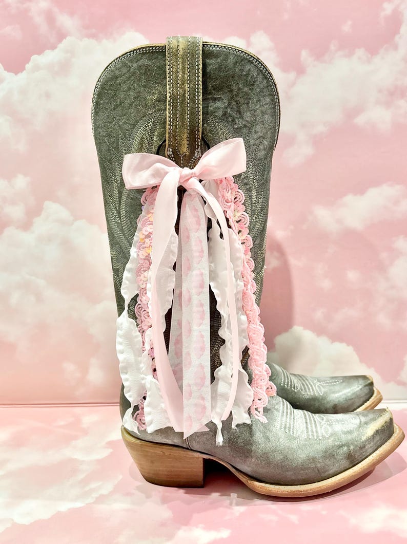 May include: A distressed grey leather cowboy boot featuring a decorative pull-on strap adorned with a multi-layered bow. The bow consists of a solid light pink ribbon, a white ruffled ribbon, and a white ribbon with a pink leopard print pattern.