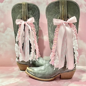 May include: A pair of distressed grey leather cowboy boots featuring a pointed toe and a low stacked heel. Each boot is adorned with a decorative light pink satin ribbon bow, accompanied by cascading white ruffled trim and pink sequined lace.