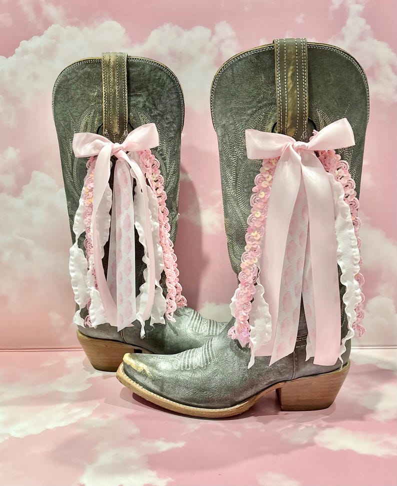 May include: A pair of distressed grey leather cowboy boots featuring a pointed toe and a low stacked heel. Each boot is adorned with a decorative light pink satin ribbon bow, accompanied by cascading white ruffled trim and pink sequined lace.