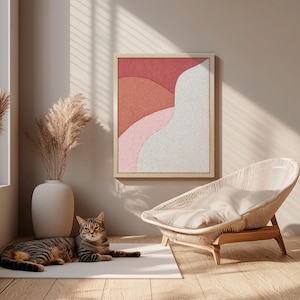 Op de afbeelding: Een abstract kunstwerk in een houten lijst toont gelaagde vormen in rood, roze en wit. Een gevlochten stoel met een crèmekleurig kussen staat in de buurt. Een tabby kat ligt op een wit tapijt.