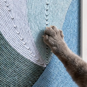 Puede incluir: Un tapiz de pared compuesto por cuerda gruesa enrollada en patrones geométricos de azul claro, azul medio y verde azulado oscuro. La cuerda está fuertemente enrollada en un diseño circular con costuras visibles, creando una superficie texturizada con una pata de gato gris azulada.