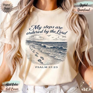 Op de afbeelding: Zacht crèmekleurig T-shirt met een marineblauwe strandscène. Het ontwerp toont voetstappen, golven en zeemeeuwen, met de tekst "My steps are ordered by the Lord" en "PSALM 37:23". Het shirt heeft een ronde hals.