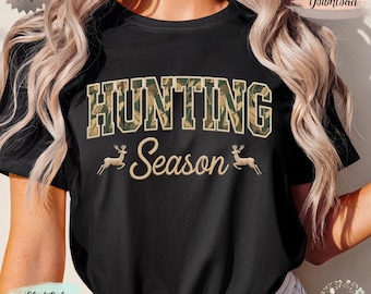 May include: Black t-shirt with the words "HUNTING Season" in a camouflage design. The word "HUNTING" is in large, block letters, and "Season" is in a cursive font. Two deer silhouettes flank the word "Season".