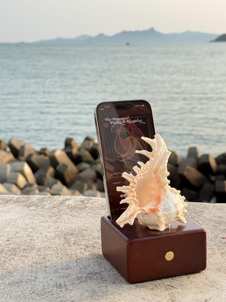 May include: A wooden phone stand with a seashell on top. A smartphone is in the stand, displaying the album art for "The Essential Perry & Kingsley".