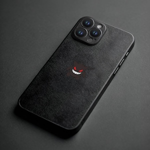 K&ouml;nnte beinhalten: Schwarze Smartphone-H&uuml;lle mit strukturierter Oberfl&auml;che. Auf der H&uuml;lle ist eine dunkelbraune Cartoon-Grafik eines l&auml;chelnden Geistes mit roten Augen und Z&auml;hnen zu sehen. Das Telefon hat eine Dreifach-Kamera.