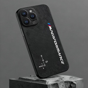 Op de afbeelding: Zwarte telefoonhoes met de tekst "M Performance" in wit en blauw. De hoes heeft een gestructureerd oppervlak en een cameramodule met meerdere lenzen. De hoes heeft ook een schakelpatroon.