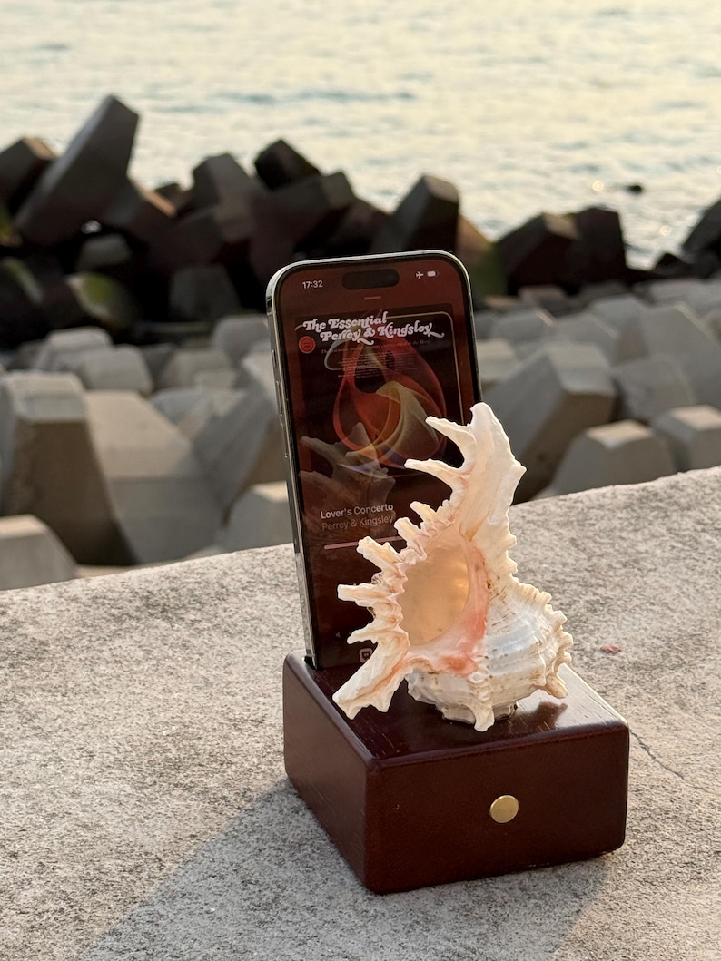 May include: A wooden phone stand with a seashell on top. The phone is playing music from the album "The Essential Percy & Kingsley".