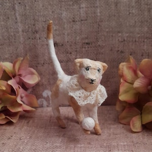 May include: A small, handcrafted cat figurine with white and tan patches, wearing a delicate white lace ruff collar around its neck. The cat is standing on three legs with its tail held high, while one front paw rests on a small, round white ball.
