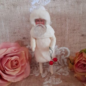 May include: A small figurine of a figure with a white fur-textured coat and matching hat, featuring a painted face with a white beard. The figure holds a thin wire ornament with two small, round, glossy red beads hanging from one hand.