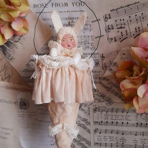 May include: A vintage-style doll ornament with a bunny-eared bonnet and a pink dress. The doll has a painted face and is adorned with lace trim. The background includes sheet music and floral accents.