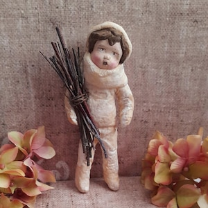 May include: A small figurine of a child with brown hair wearing a cream-colored hooded outfit and a matching scarf. The figure holds a bundle of thin, dark brown twigs tied together with wire. The face features painted eyes, nose, and mouth.