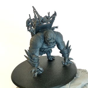 May include: A gray plastic miniature of a large, monstrous creature with a spiked tail and claws. The creature is carrying a small, wooden structure on its back. The miniature is on a black circular base.