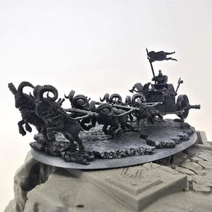 May include: A detailed miniature model of a chariot pulled by several rams. The chariot has a driver and a flag. The model is dark gray and sits on a rocky base.