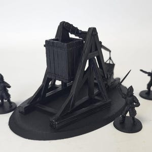 Könnte beinhalten: Schwarzes Miniaturmodell eines Trebuchets mit drei kleinen Soldatenfiguren. Das Trebuchet hat einen Holzeimer und einen komplexen Rahmen. Die Soldaten stehen auf kleinen kreisförmigen Sockeln, vor weißem Hintergrund.