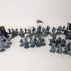Puede incluir: Colección de figuras en miniatura grises que representan una escena de batalla medieval. El conjunto incluye soldados a pie y a caballo, máquinas de asedio y banderas, todo dispuesto sobre una superficie blanca.