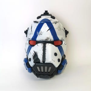 May include: A white and blue helmet with a distressed finish. The helmet features a blue stripe, red eye lenses, and black accents. The design includes a black face guard and blue side details.