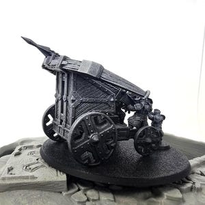 May include: A black miniature trebuchet model with two figures. The model features a detailed wooden design, large wheels, and a launching arm. The figures appear to be in the process of operating the siege weapon. The model is set on a black base.
