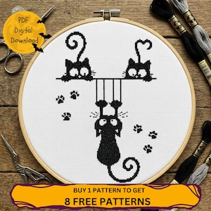 May include: A cross-stitch pattern in a wooden embroidery hoop featuring three black cats on white fabric. Two cats sit on a horizontal line while the third hangs below by its paws. The design includes small paw prints and a yellow circular graphic.