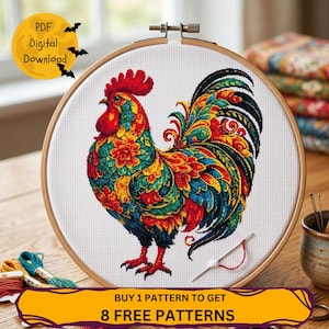 May include: A round wooden embroidery hoop containing white fabric with a cross-stitch design of a rooster. The rooster features vibrant red, yellow, green, and blue floral patterns. A needle with red thread rests on the fabric surface.