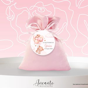 K&ouml;nnte beinhalten: Ein rosa Gastgeschenkt&uuml;te, gebunden mit einem Band, mit einem runden Anh&auml;nger mit einer Teddyb&auml;r- und Ballonillustration. Der Anh&auml;nger lautet "Il Battesimo di Gaia 15 Giugno 2025". Der Hintergrund ist hellrosa mit wei&szlig;en floralen Linien.