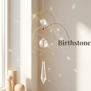 May include: A gold-toned metal suncatcher featuring two interlocking circular hoops, a clear faceted crystal bead, a small red birthstone, a larger clear crystal sphere, and a clear crystal prism pendant hanging from a delicate gold chain.