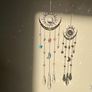 Puede incluir: Dos atrapasoles de temática celestial cuelgan de cadenas. Uno presenta un diseño de sol y media luna con cristales colgantes, cuentas y una estrella azul. El otro tiene un diseño similar con una esfera oscura en el centro del sol.