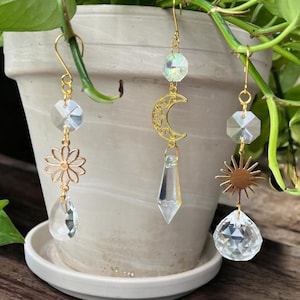 Pode incluir: Três brincos dourados com desenhos celestiais e florais. Um apresenta uma margarida, outro uma lua crescente e o terceiro um sol. Cada brinco tem detalhes de cristal transparente e é suspenso por um gancho dourado.