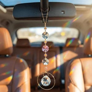 Peut inclure: Un ornement de voiture suspendu composé d'une chaîne dorée avec de petites fleurs marguerites roses et blanches, trois cristaux facettés octogonaux transparents et roses, et un grand cristal sphérique suspendu dans un anneau doré.