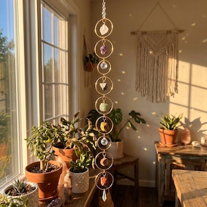 May include: A hanging crystal suncatcher with gold-colored rings and various gemstones. The suncatcher is suspended in front of a window, with sunlight illuminating the crystals and creating rainbows. The gemstones include clear, purple, black, green, and orange stones.
