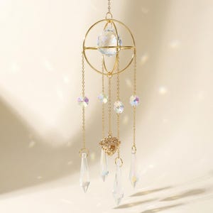 May include: A gold-colored suncatcher with a large crystal ball at the center, surrounded by a circular frame. Several smaller iridescent crystals and teardrop-shaped prisms hang below, connected by delicate chains.