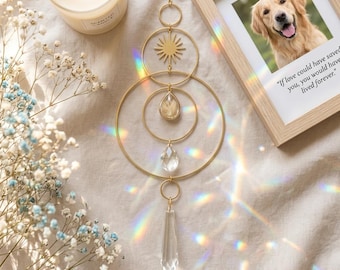 May include: A gold-toned metal suncatcher featuring three concentric rings, a sunburst emblem, a teardrop-shaped crystal, and a clear glass prism pendant. The piece hangs vertically, casting rainbow light refractions across the surrounding area.