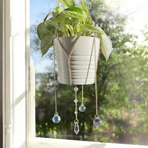 Puede incluir: Una maceta colgante de cerámica blanca con una planta verde y frondosa, suspendida por cadenas doradas. Las cadenas están adornadas con detalles de cristal transparente e iridiscente. La maceta cuelga frente a una ventana.