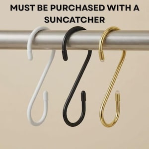 May include: Three S-shaped hooks in white, black, and gold hang from a silver bar. The hooks are designed for hanging items. Text at the top reads "MUST BE PURCHASED WITH A SUNCATCHER".