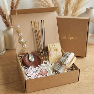 May include: A cardboard box containing spiritual items. It includes incense sticks, a necklace, a wooden bowl, a bundle of sage, and a crystal suncatcher. The box also has stickers with inspirational quotes. The lid is printed with the words "Sun Aura".