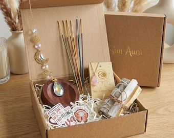 May include: A cardboard box containing spiritual items. It includes incense sticks, a necklace, a wooden bowl, a bundle of sage, and a crystal suncatcher. The box also has stickers with inspirational quotes. The lid is printed with the words "Sun Aura".
