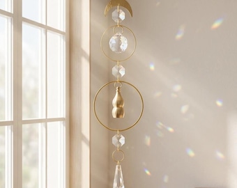May include: A hanging suncatcher featuring a gold-toned crescent moon, two thin circular metal rings, and a central brass gourd-shaped charm. The design includes clear faceted crystal beads and a pointed crystal prism, casting light reflections.